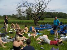 People having lunch in the grass with canoes in the background. Photo. 